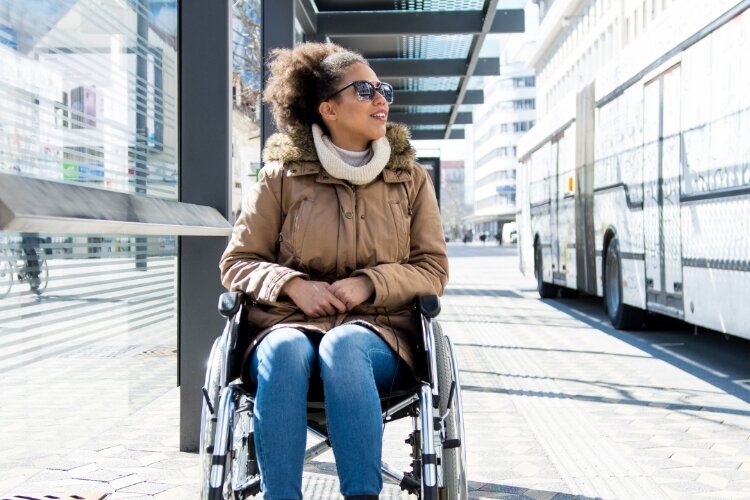 Photo shows a woman in a wheelchair at a bus stop.