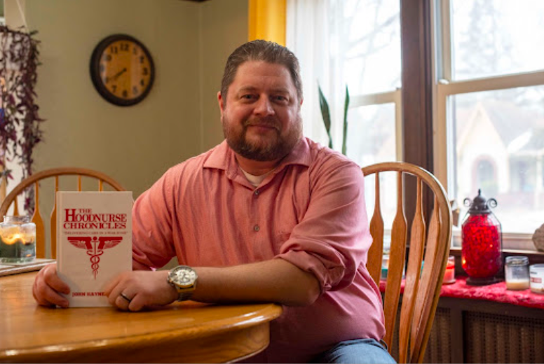 John Haynes sits inside his dining room, holding his first physical copy of 'The Hoodnurse Chronicles: Delivering care into a war zone.'